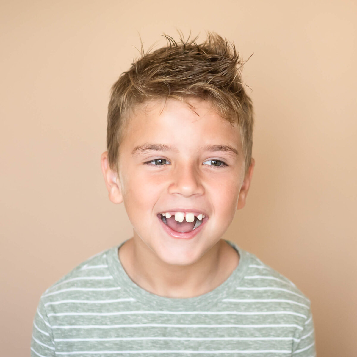children's studio portrait of young boy laughing in sage and white striped shirt with a tan background