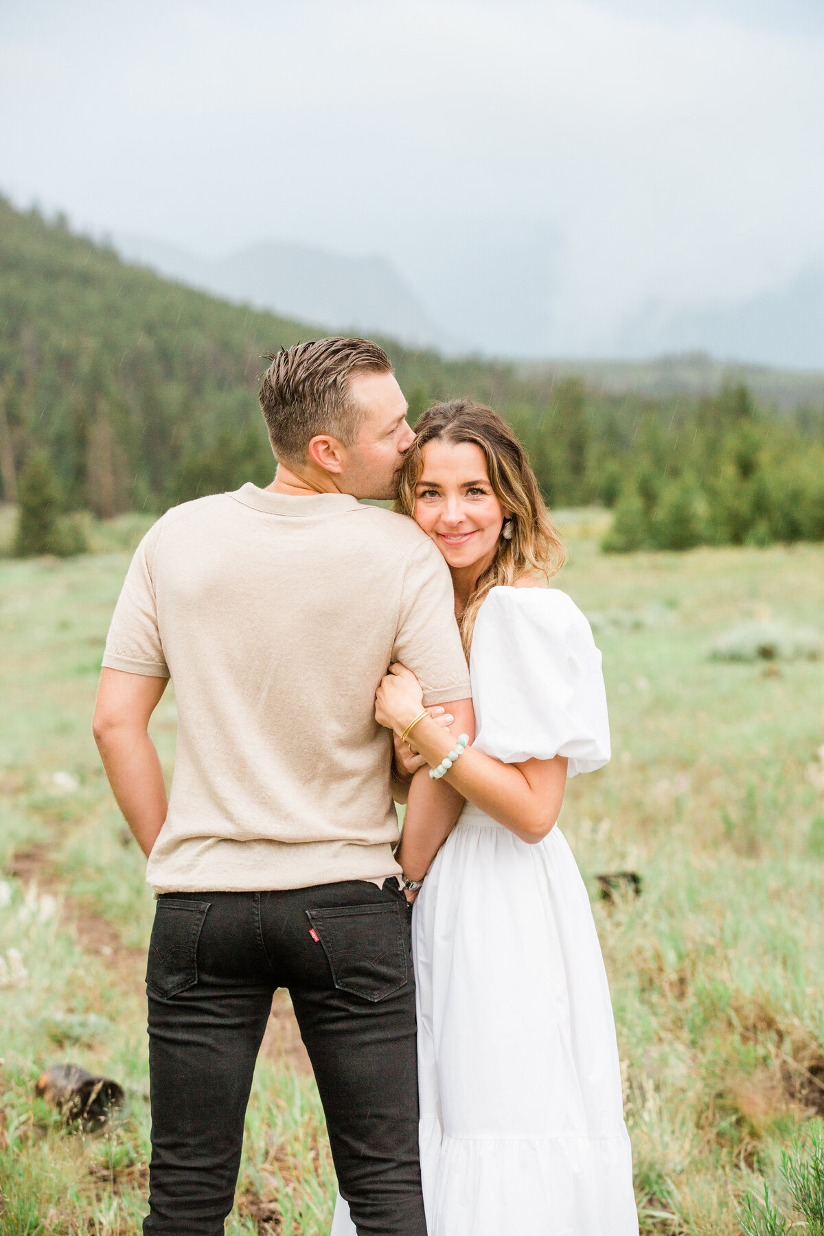 This couple is in a meadow on Swan Mountain. The husband is kissing his wifes forehead as she looks and smiles at the camera