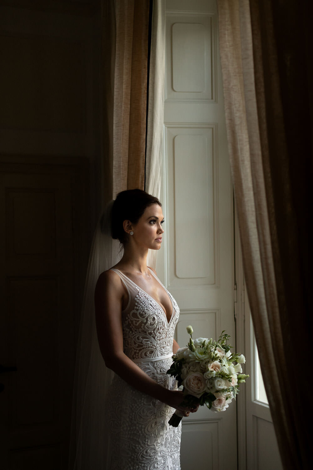 Bride holding floral bouquet in white lace wedding dress looking out window in lake Bride holding floral bouquet in white lace wedding dress looking out window in lake Como Wedding Italy Wedding Italy
