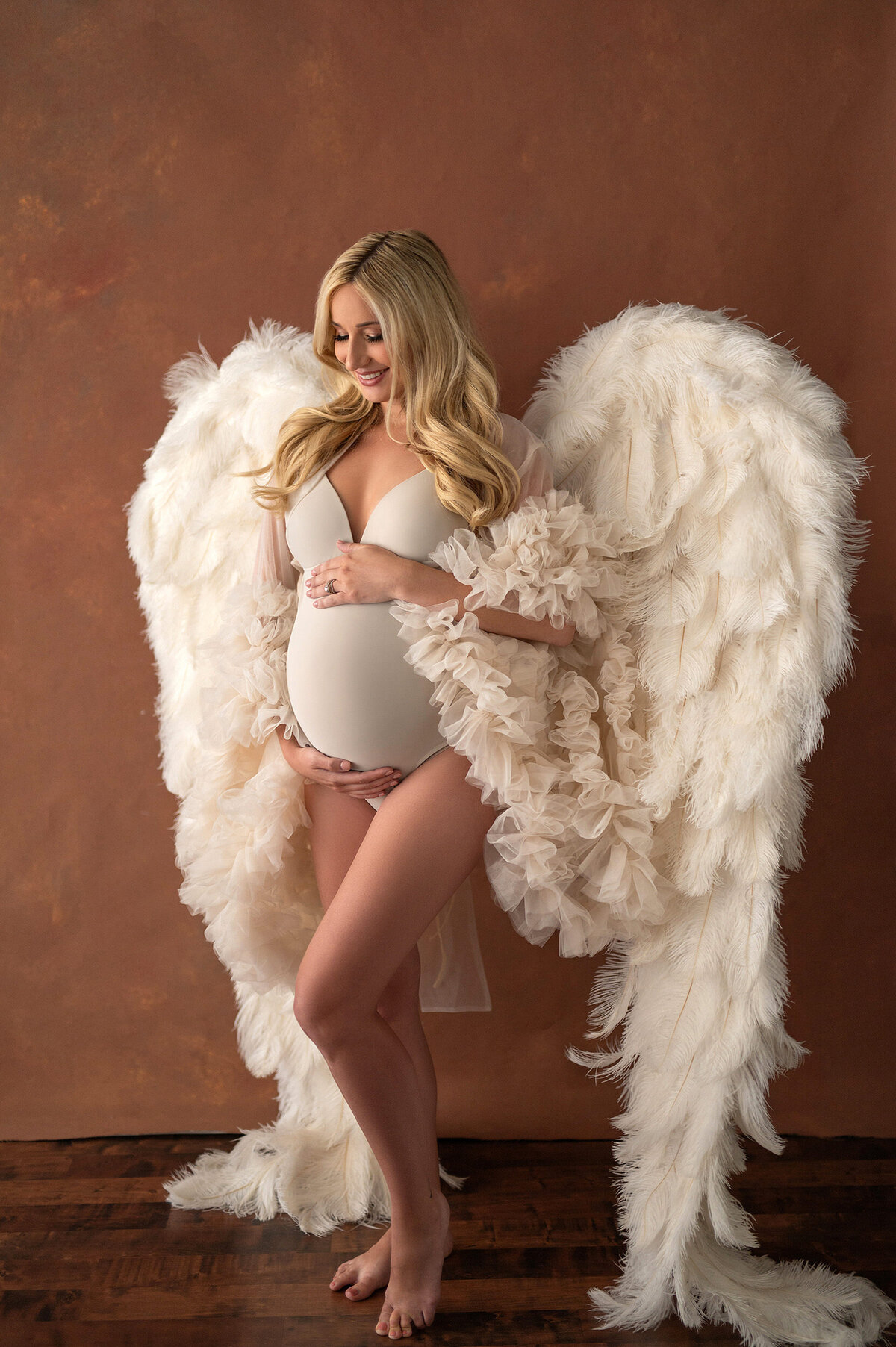 Full length image of a pregnant woman in an ivory bodysuit and floor length angel wings cradling her belly in our Waukesha Wisconsin studio .