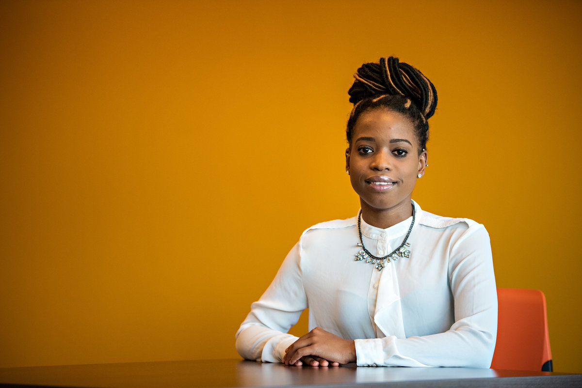 A young professional woman sits at a desk in front of an orange wall.