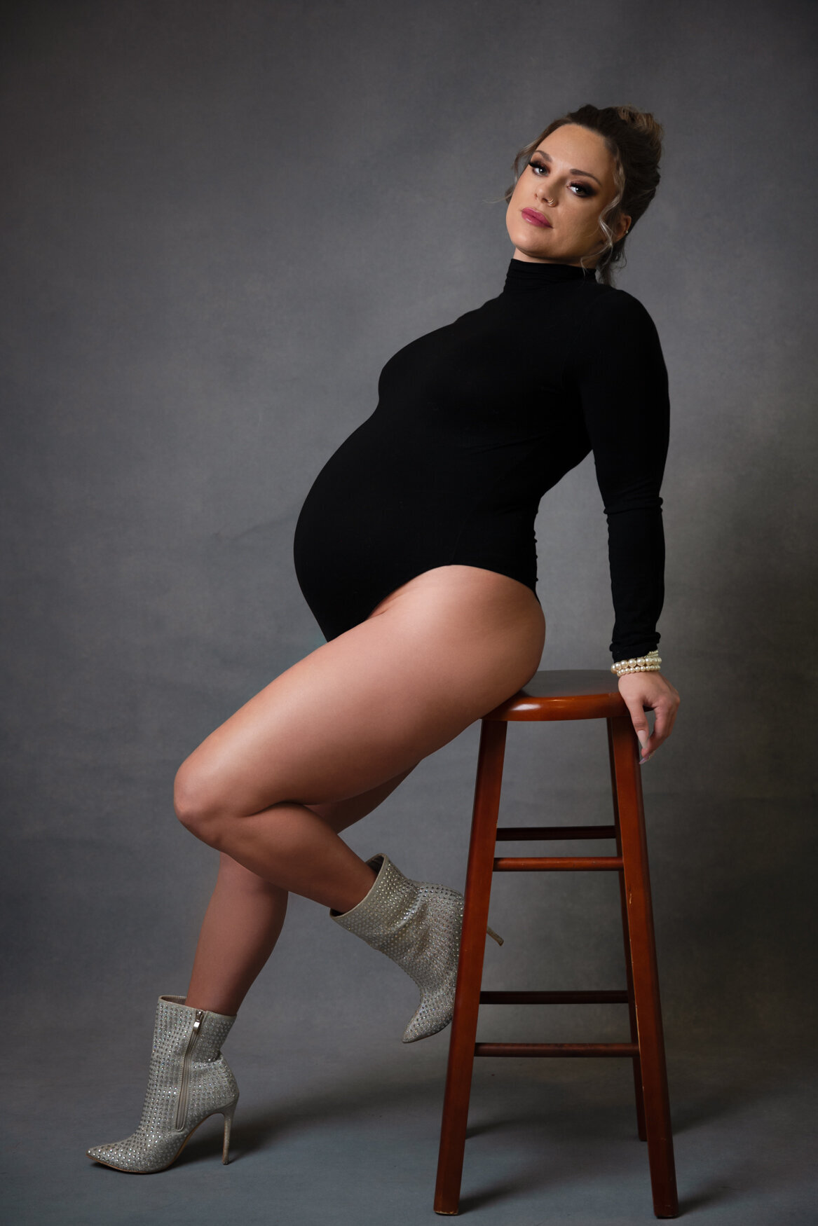 A woman wearing a black bodysuit and silver boots posing on a stool showing her  pregnant belly