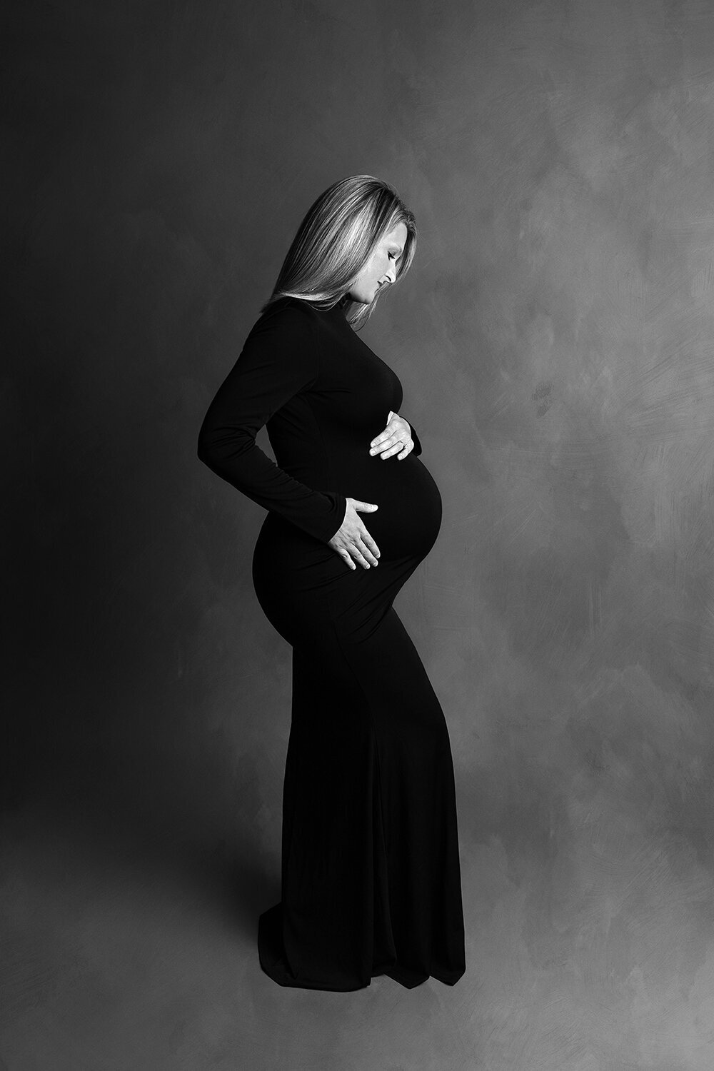 B&W image of a pregnant woman in a black maternity gown with hands on her bump
