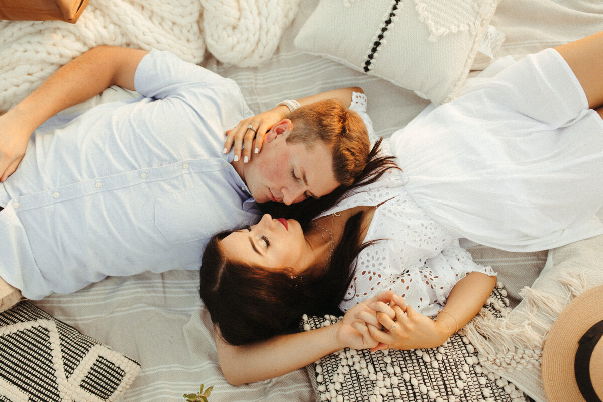 Couple laying down on a blanket face to face and holding hands with pillows around them