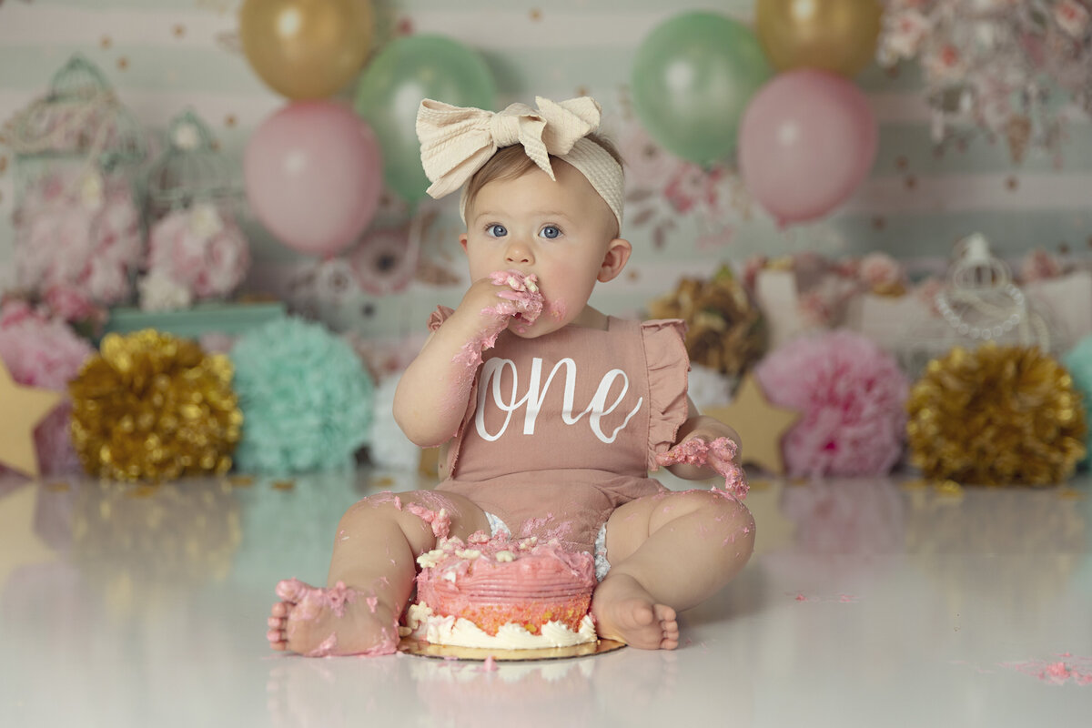 A toddler girl in a big bow and pink onesie sits for a New Jersey Cake Smash Photographer covered in cake