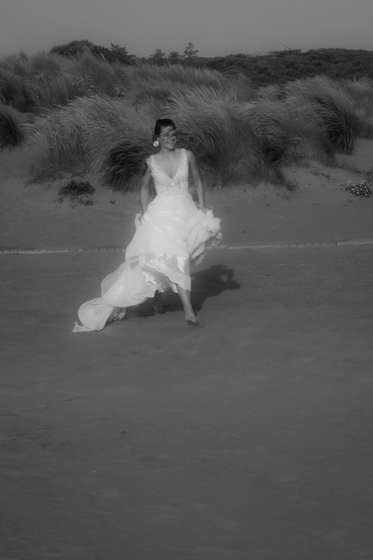 Mit wehendem Kleid rennt die strahlende Braut vor einer bewachsenen Düne über den Strand.