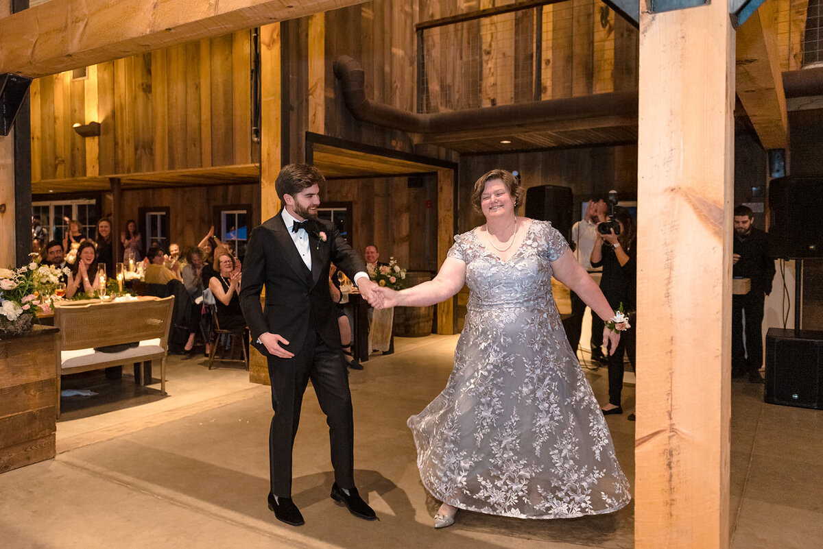 A mother and groom dancing, with the mother in a lace dress, in the warm, rustic interior of a barn.