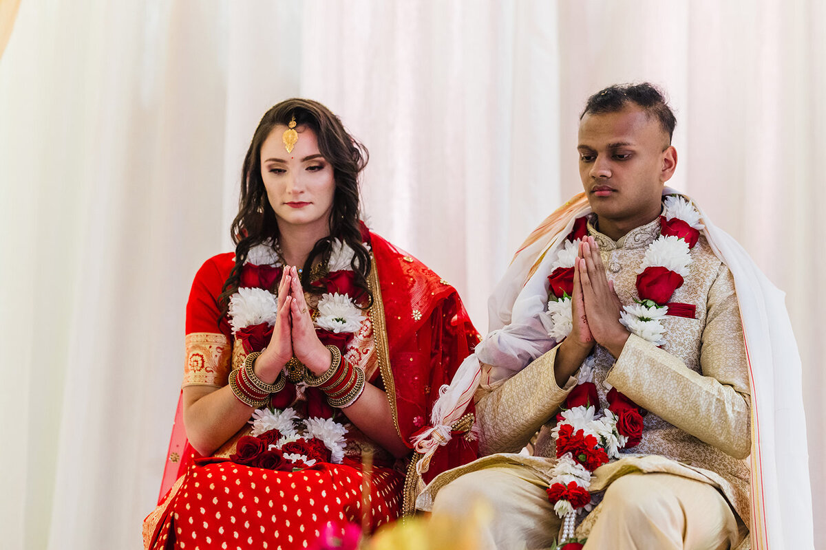 A bride and groom in traditional Indian wedding attire sitting with folded hands in a prayerful pose during a ceremony.