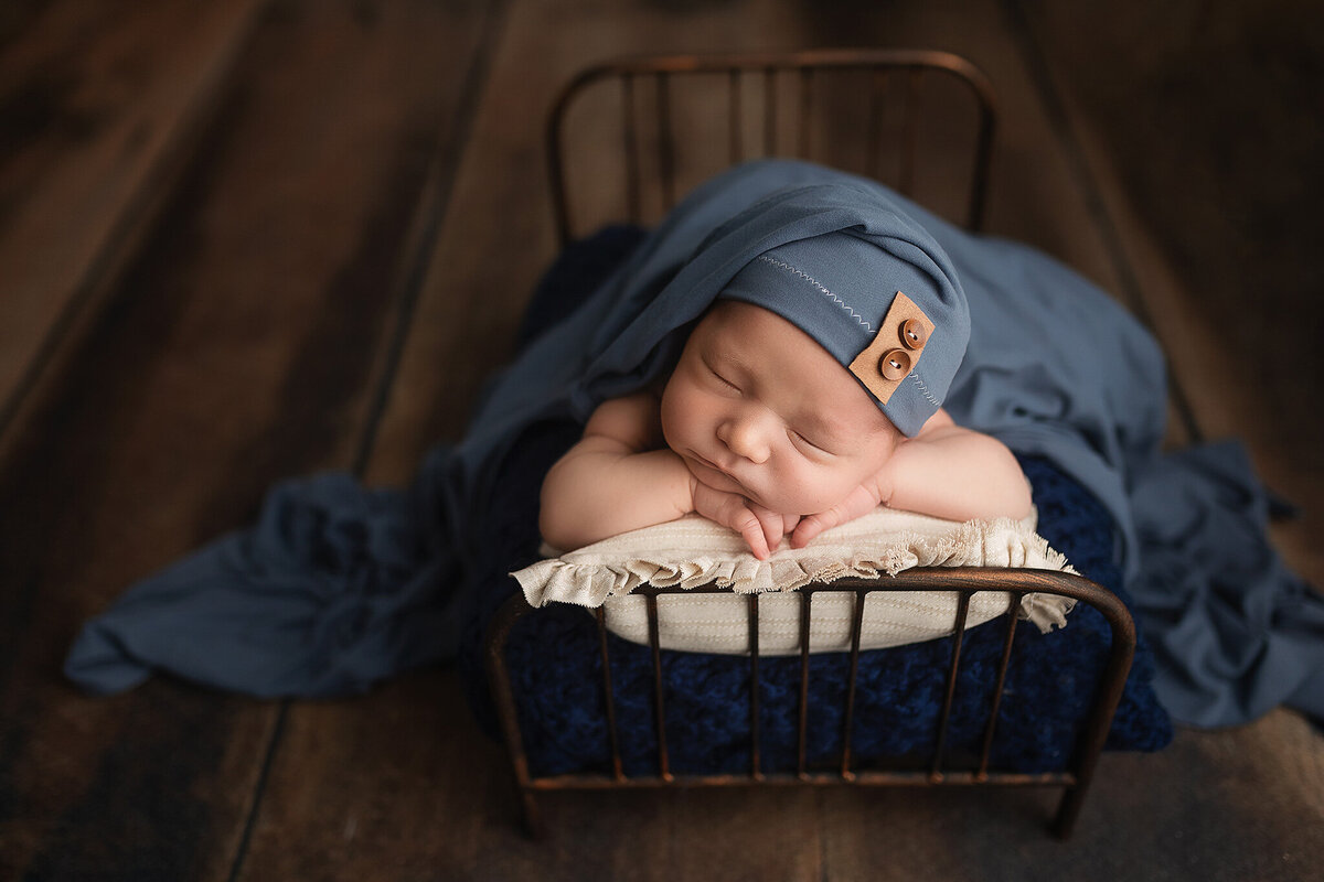 Baby boy wearing a blue sleepy cap while posed on newborn bed prop.