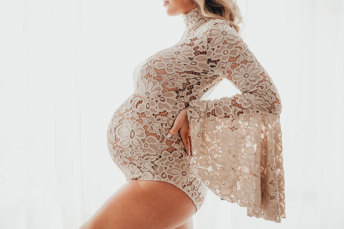mother holds her belly against all white backdrop while holding her arched back, wearing winged arm bodysuit.