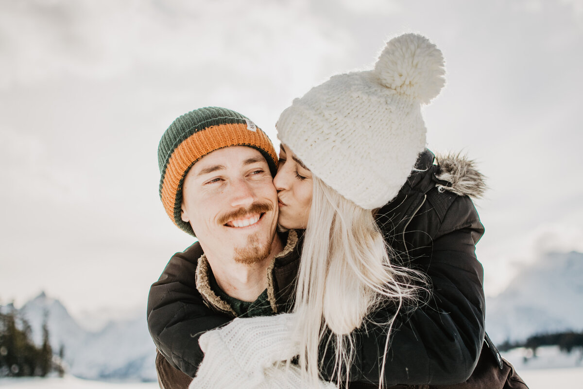 woman behind her fiance kissing his cheek as he smiles during their winter Jackson Hole engagement session with jackson hole photographers