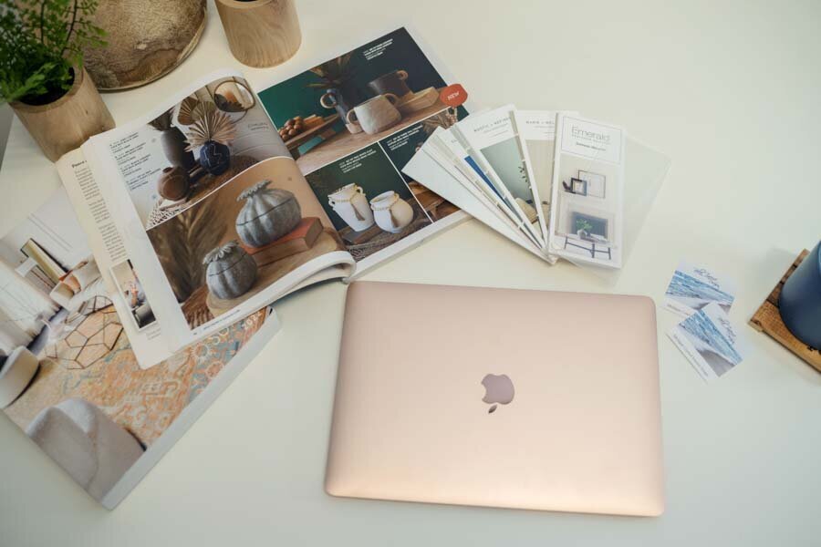 A rose gold laptop beside a spread of design magazines and paint samples on a white table, with greenery around.