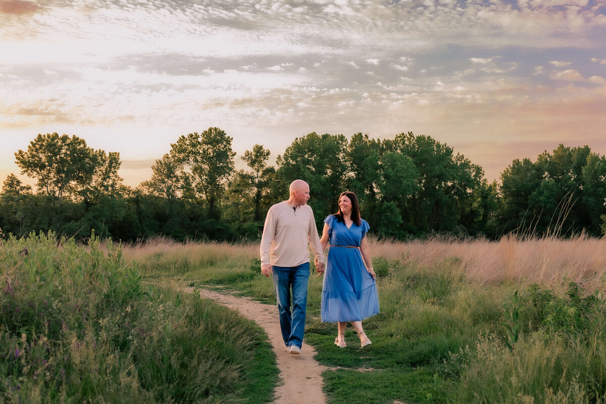 A Couple walking hand in hand on a dirt path sounded by green tall grass and a colorful cloudy sky at sunset.