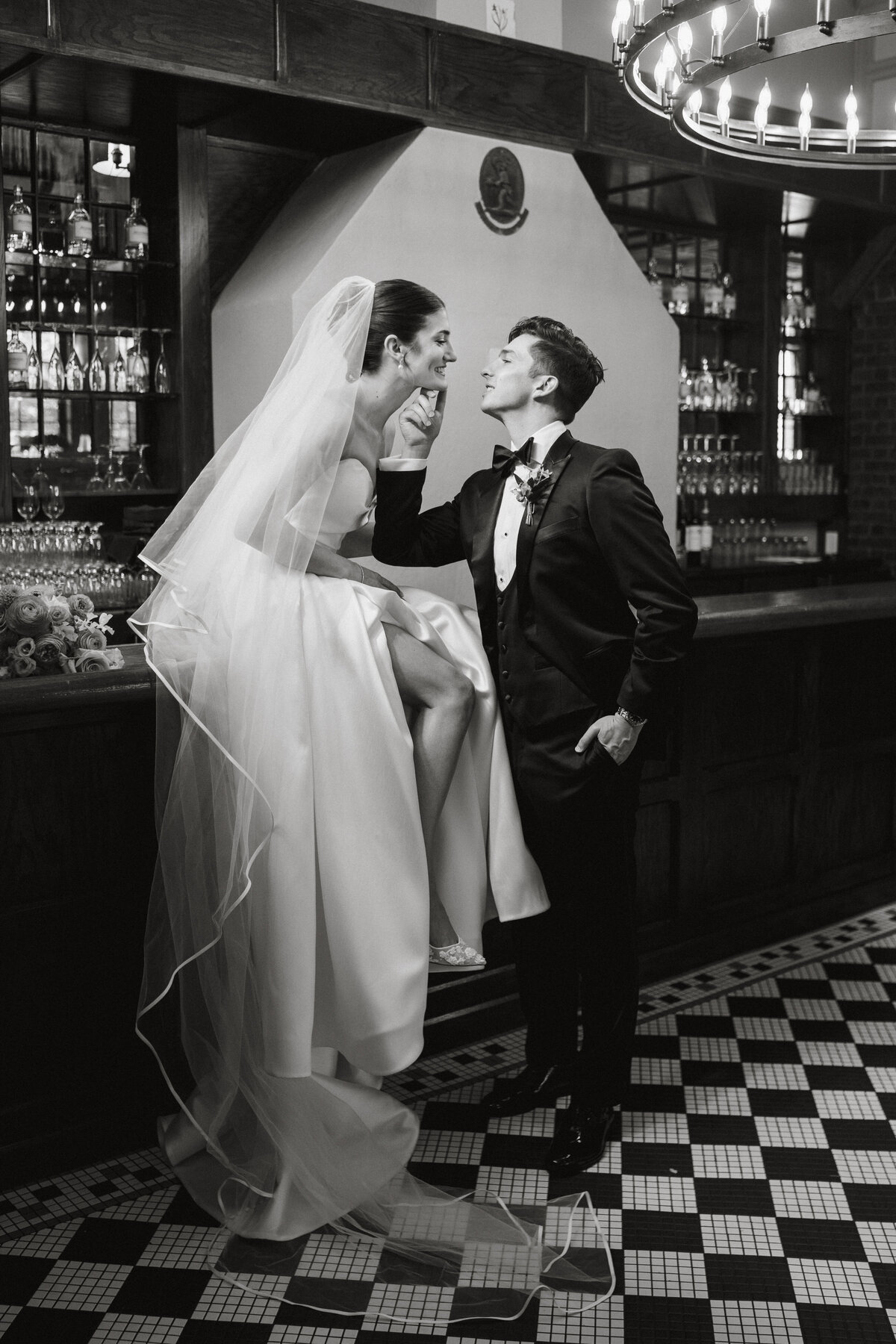 Woman sitting on bar in wedding dress while groom kisses her
