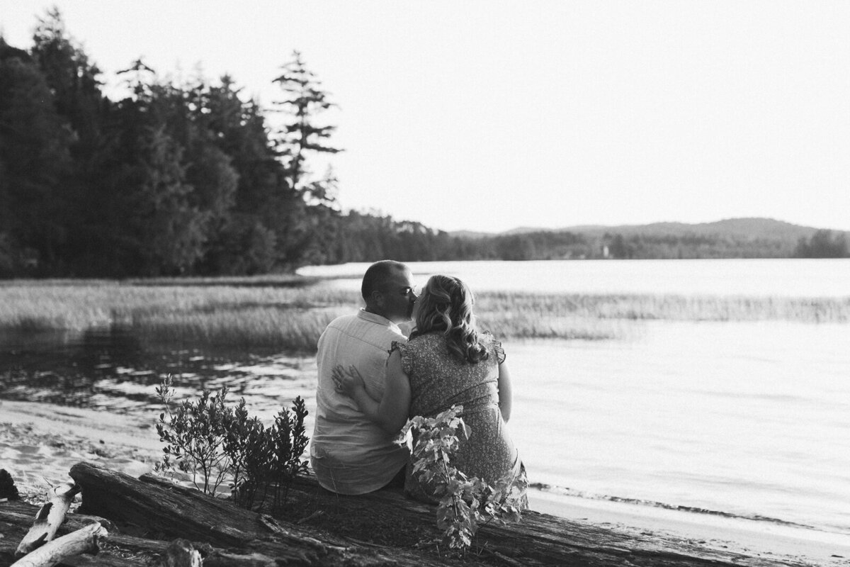 couple faces the water  sitting on a log and kisses