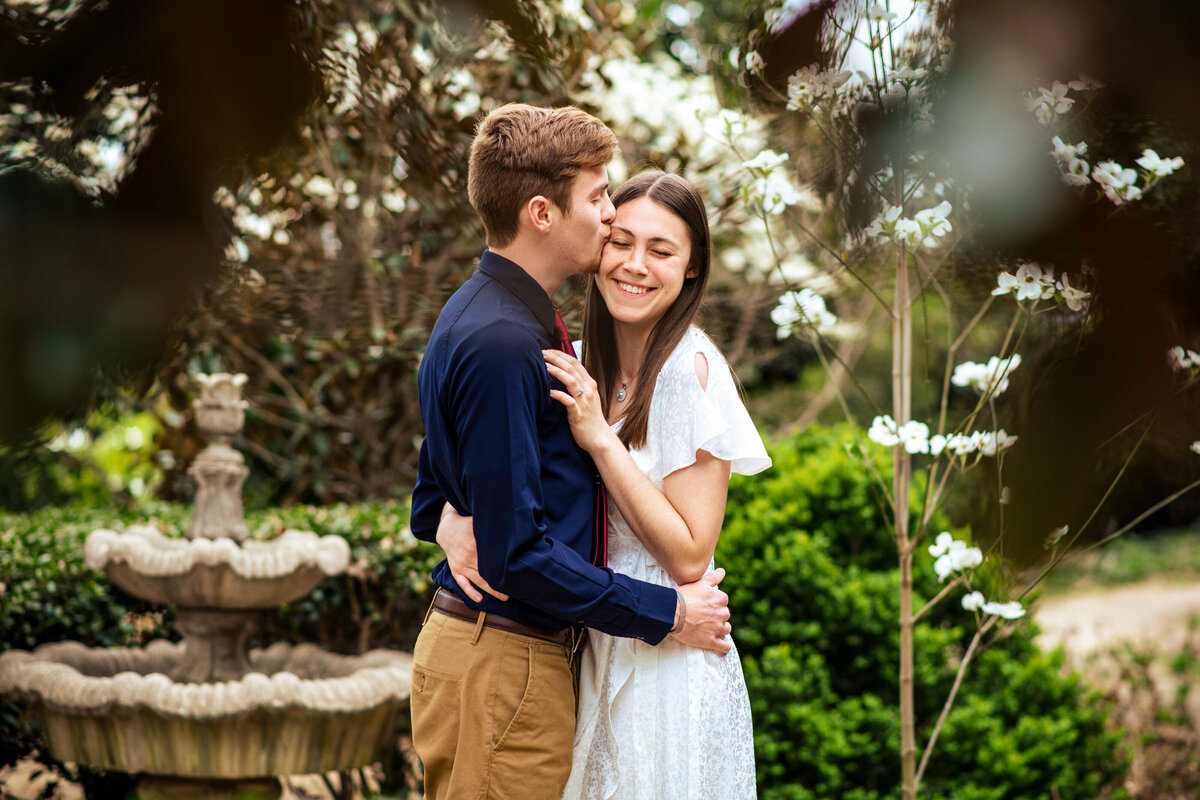 Female-giggling-with-her-eyes-closed-as-her-fiance-whispers-in-her-ear-amid-greenery-and-white-spring-flowers-at-the-McGill-Rose-Garden-in-Charlotte