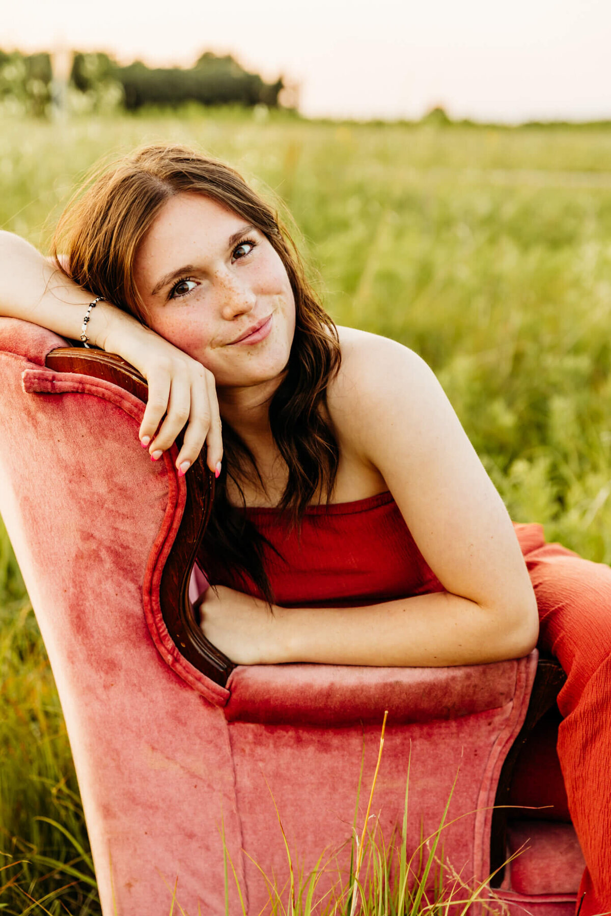 brown hair high school girl resting on a pink velvet chair in a field near Oshkosh