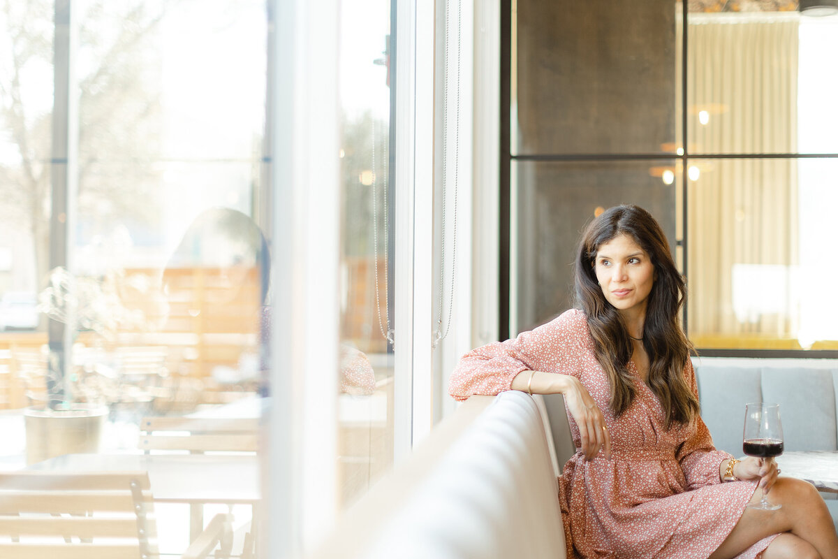 Professional headshot of a Dallas/Fort Worth business owner sitting down as she looks out the window while holding a glass of wine.