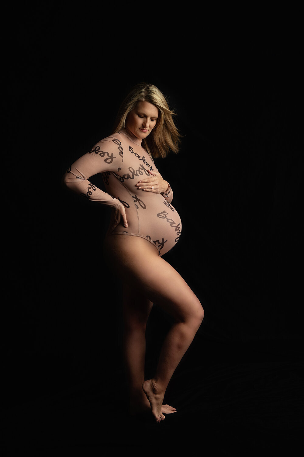 A blonde pregnant woman pops a leg while standing in a studio in a tan maternity one-piece