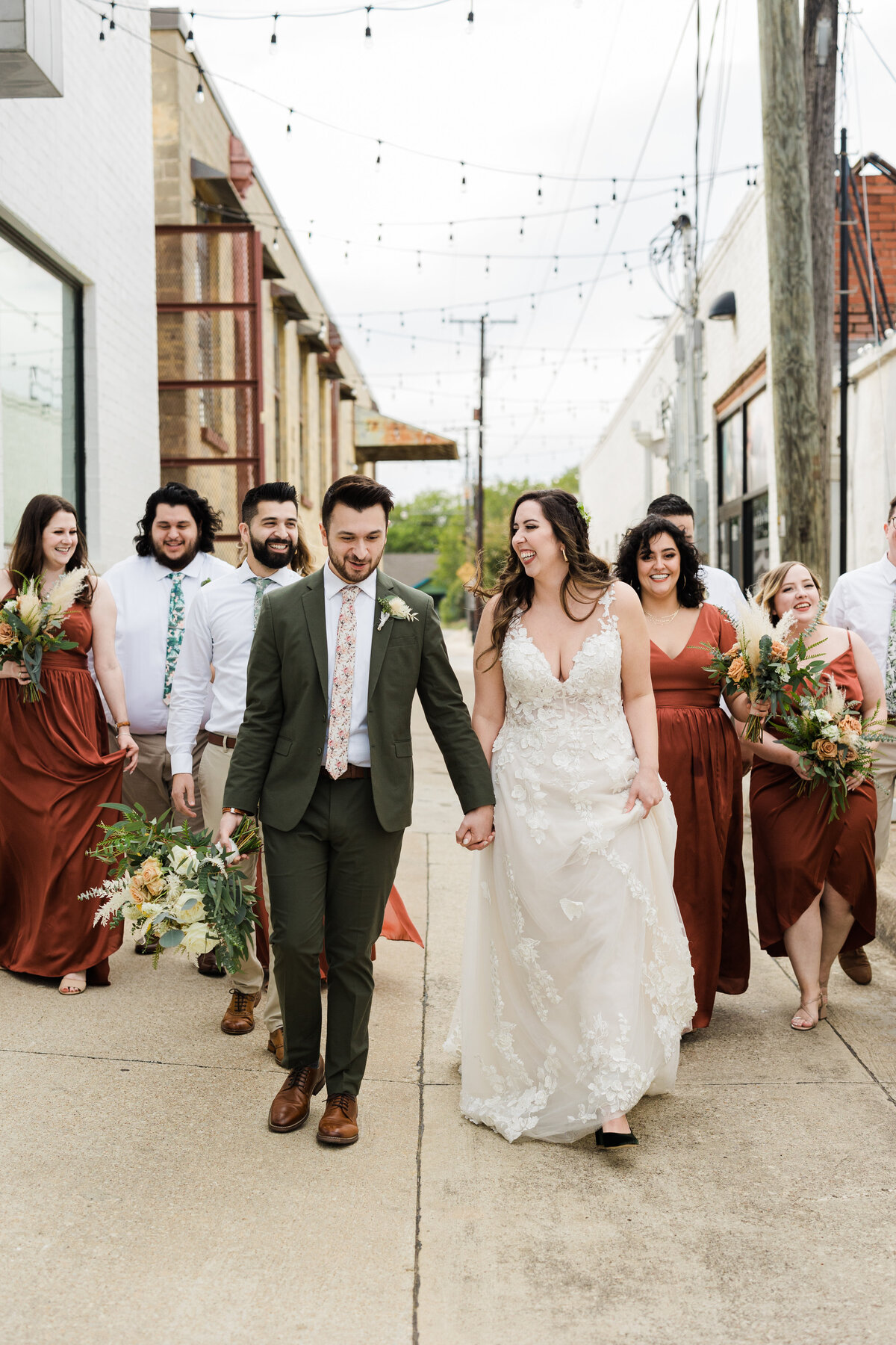 A walking shot of a bride, groom, and their whole wedding party after their wedding ceremony at The 4 Eleven in Fort Worth, Texas. The bride is on the right and is wearing a long, intricate, white dress with a hairpiece. The groom is on the left and is wearing a dadrk green suit with a floral tie and boutonniere. Their wedding party flanks them on either side; the groomsmen are wearing white dress shirts, khaki dress pants, and varying floral ties while the bridesmaids are wearing dark orange dresses with bouquets.