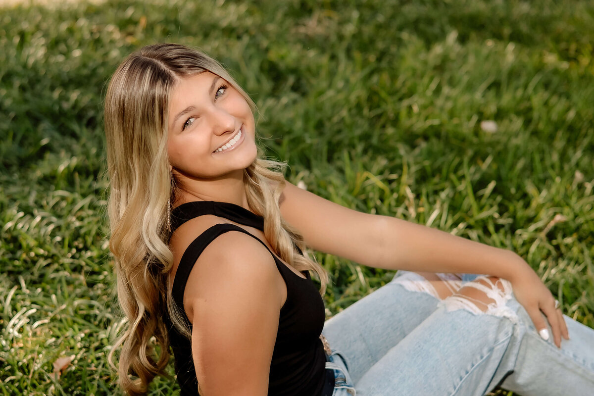 a high school senior girl sitting in the grass looking over her shoulder back at the camera