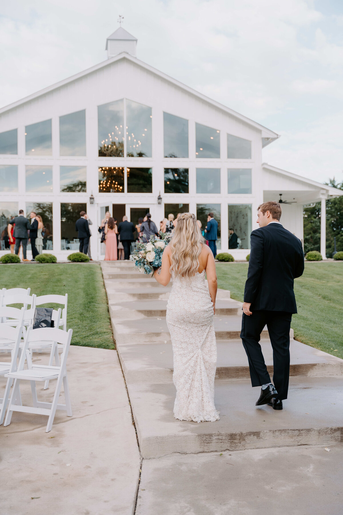A bride and groom walk towards a modern white chapel with large windows and a crowd gathered outside. The scene feels celebratory and elegant.