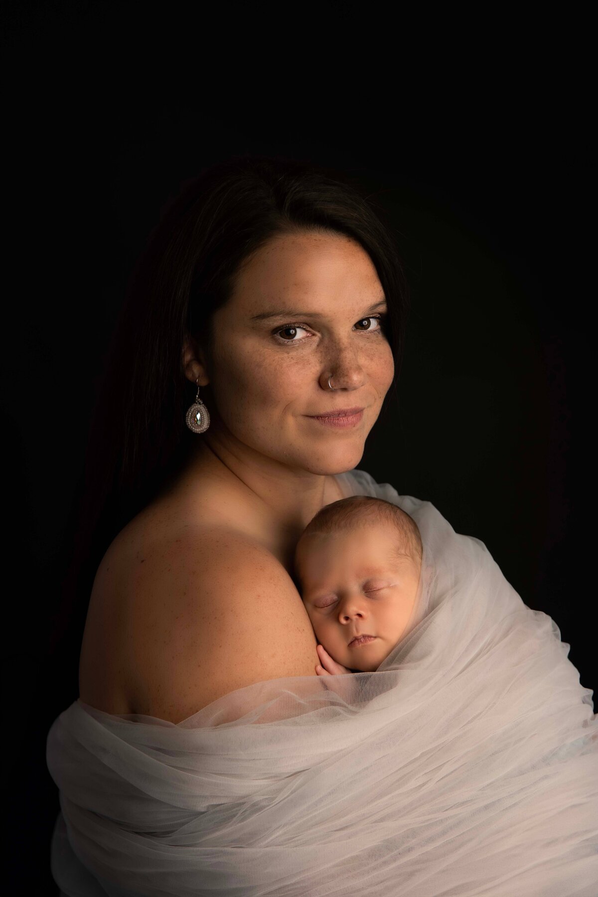A gorgeous new mom poses for a newborn portrait with her baby boy. He is sleeping peacefully on her chest, and the two of them are wrapped in grey tulle. They are snuggling skin-to-skin as the baby sleeps peacefully on his mom and she glances at the camera.
