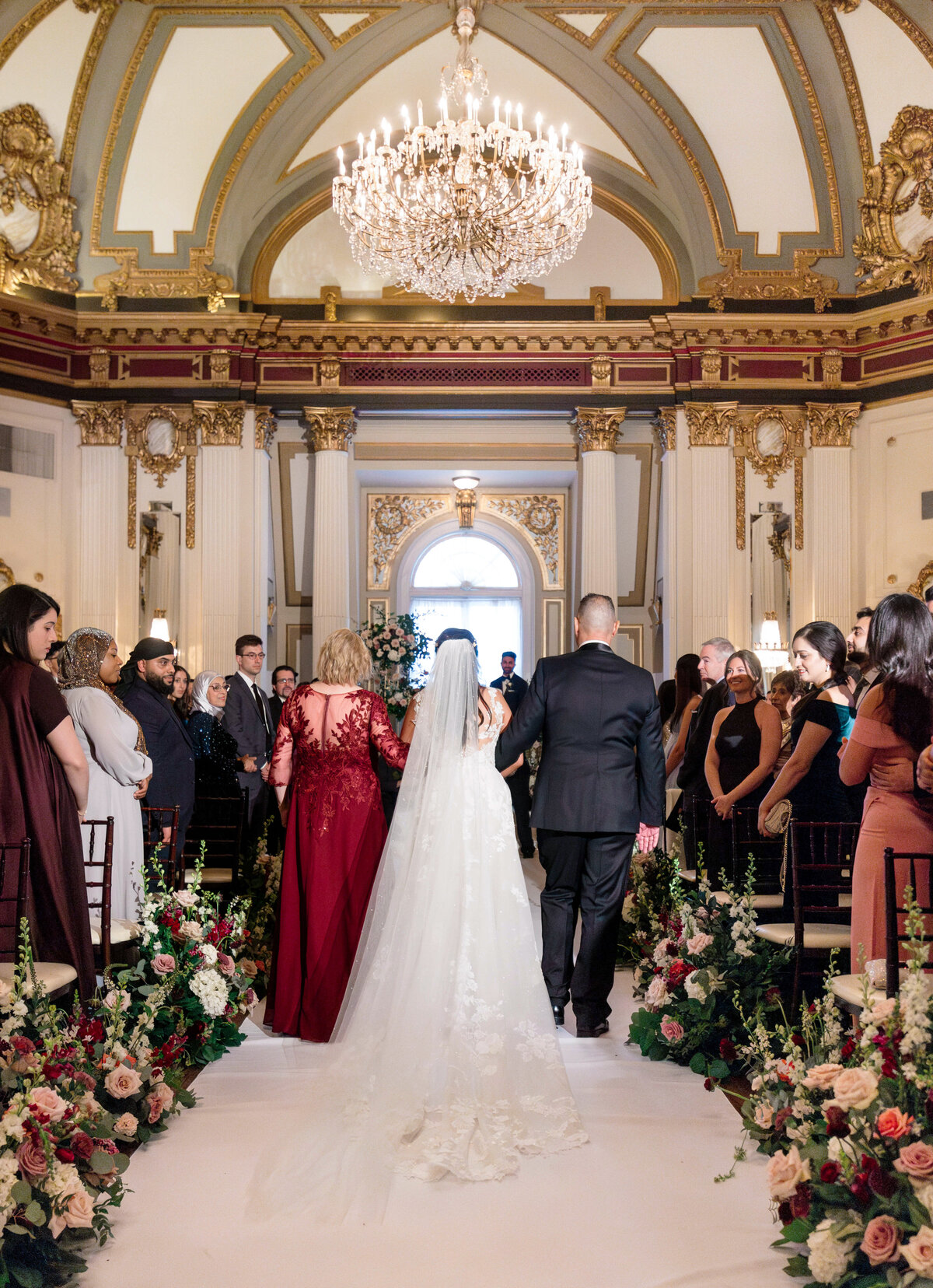 A bride walks down the aisle with two people in an ornate hall adorned with a grand chandelier and floral decorations. Guests stand on either side, observing the wedding ceremony.