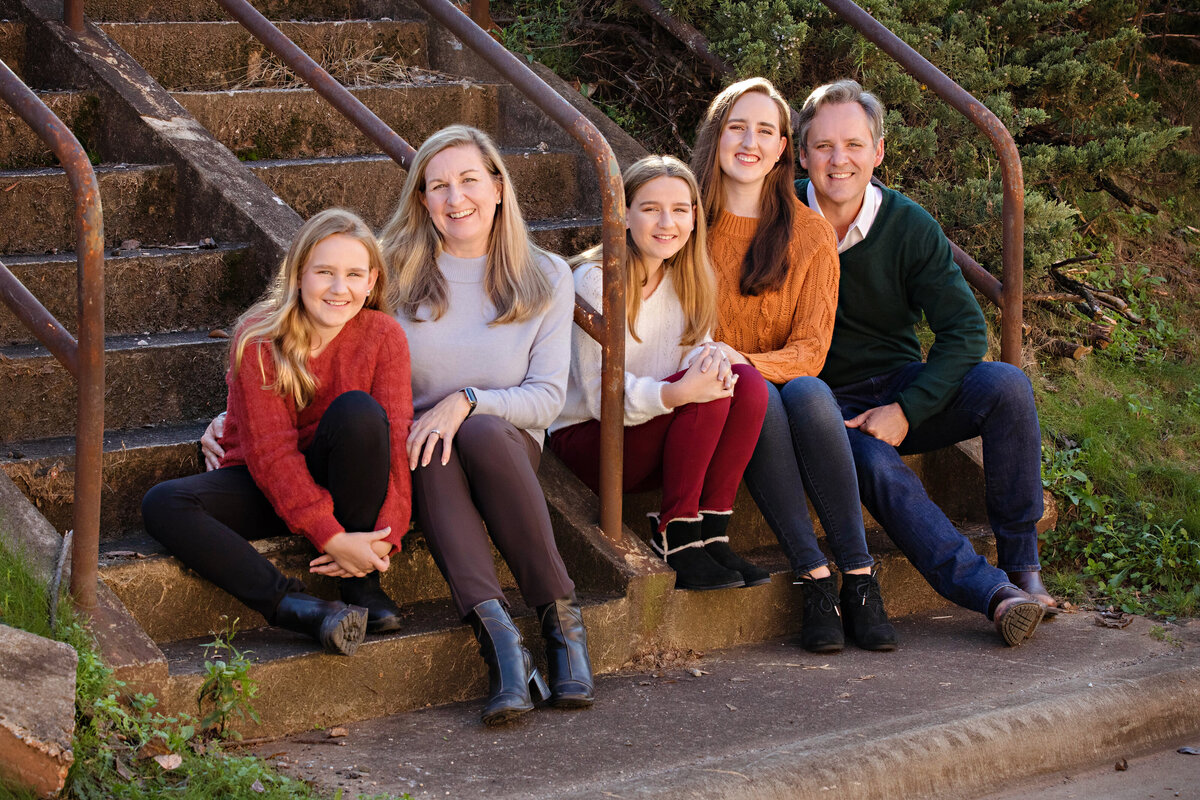 Family portrait in a multi-color autumn sweater theme that matches perfect with the urban surroundings of Camp North End.  Family of five is sitting on a concrete staircase with rusted railings that match the color tones of the rust and orange sweaters or brown pants.