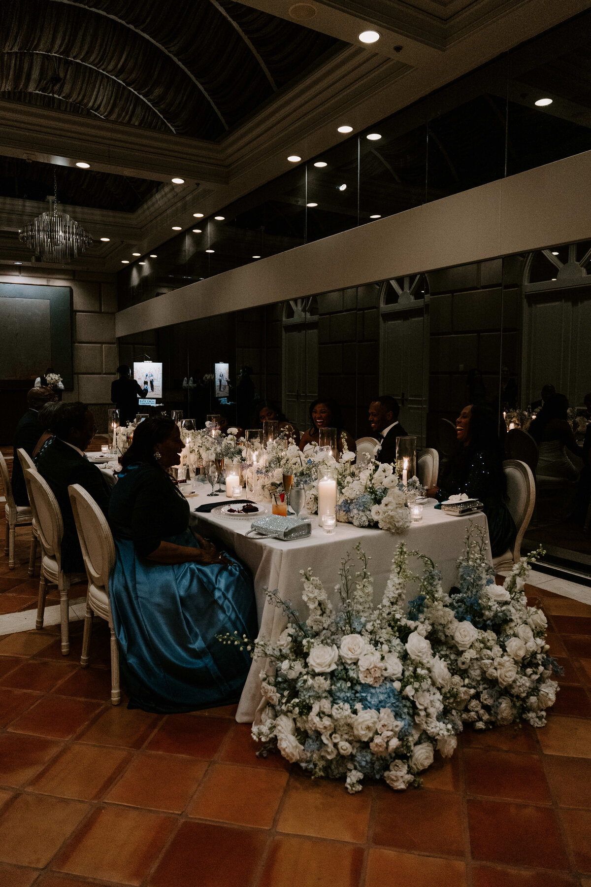 Elegant dining setup in a dimly lit room with mirror walls. A long table is adorned with white flowers, candles, and guests in formal attire, evoking a sophisticated ambiance.