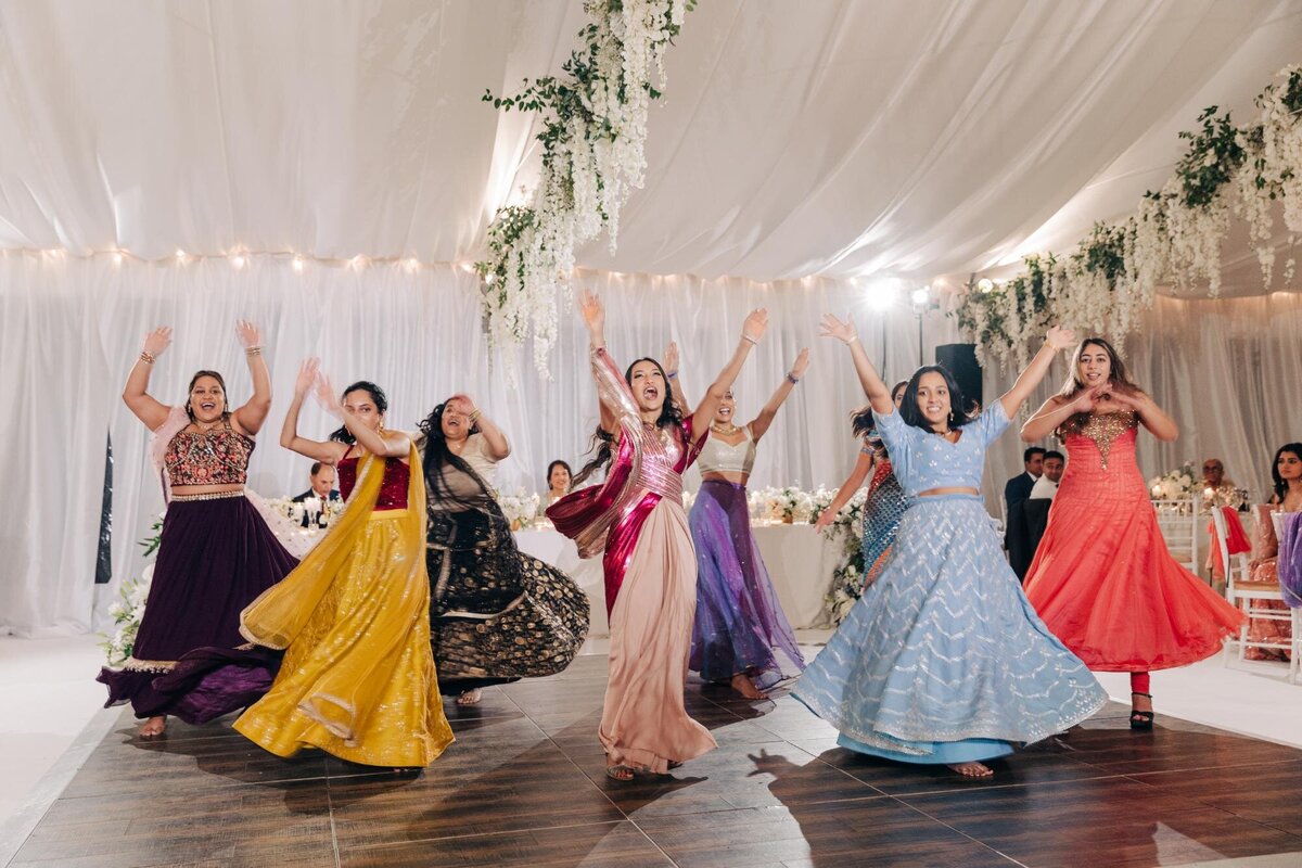 Group of women joyfully dancing in colorful traditional outfits at a festive indoor event with floral decorations.