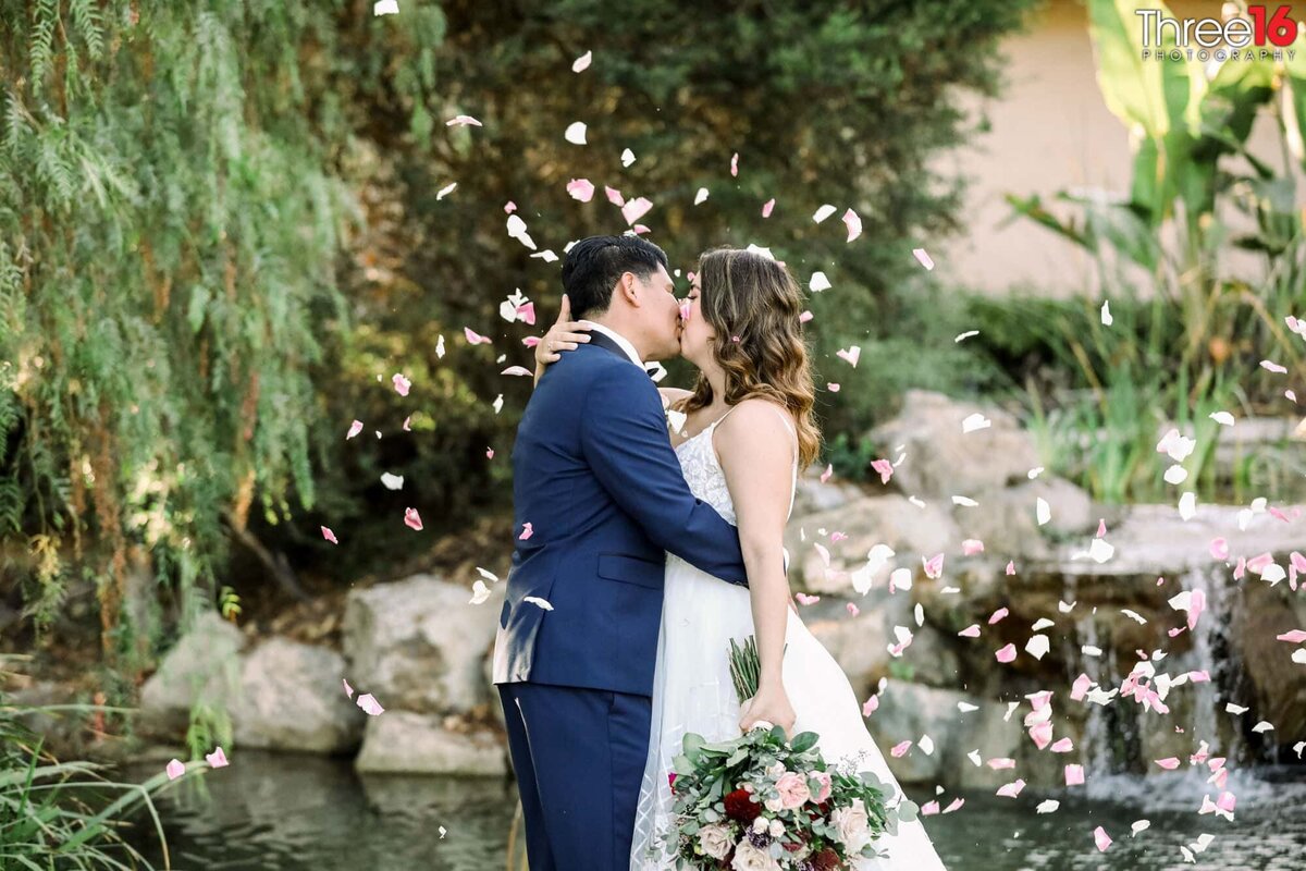 Bride and Groom share a romantic kiss in front of the pond and waterfall with rose pedals raining down on them
