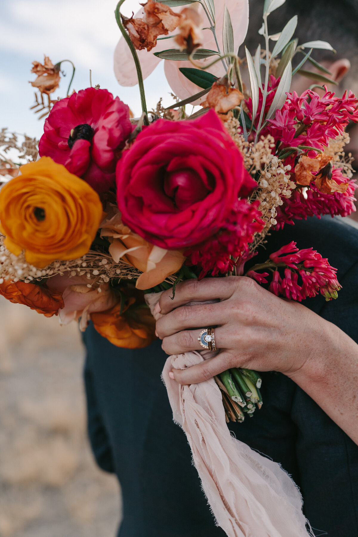 A colorful photograph of Maggie and Caleb on their wedding day at La Tierra in Marfa, Texas. The photograph is a close-up of the bride holding her bouquet behind the back of the groom. The wedding ring is sparking. The ring is soft gold with a diamond in the center. The wedding band is a row of diamonds on one side and a half circle of gray stone and diamonds on the other side. The bouquet the bride is holding is magenta, orange and yellow and blue-ish green eucalyptus with a light pink soft ribbon flowing down. Wedding photography by Stacie McChesney/Vitae Weddings.