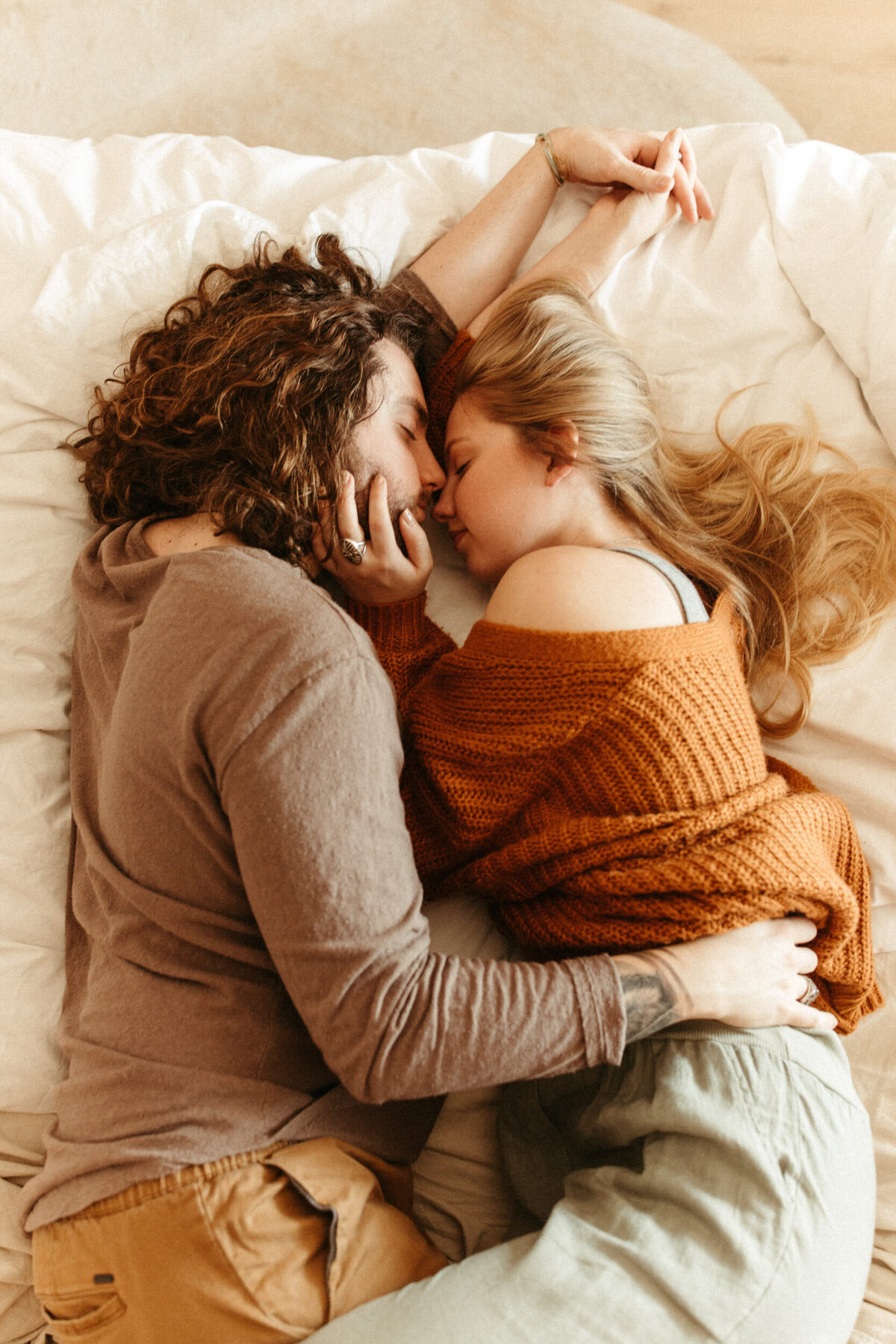 A couple wearing cozy outfits is snuggling and laying on a bed holding hands.
