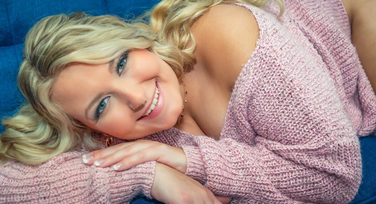 A blond woman in a blush pink sweater smiles at the camera as she poses on a blue couch for her boudoir photoshoot.