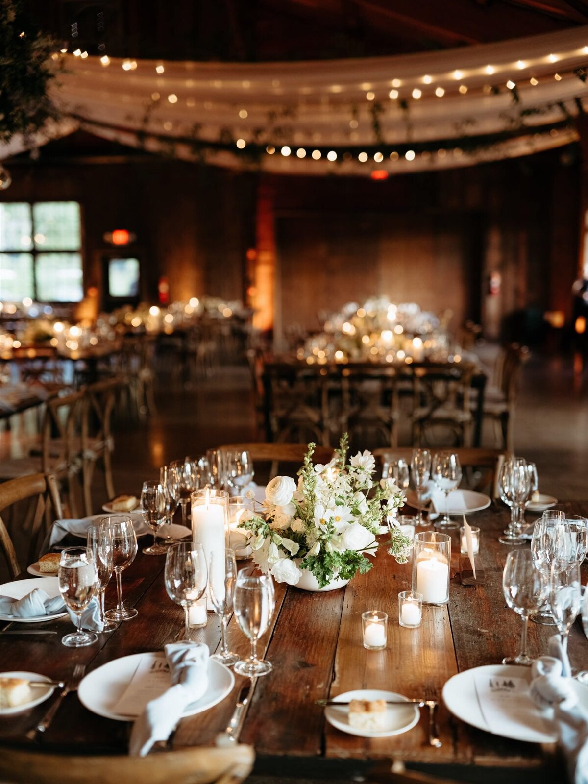 Cedar Lakes Estate wedding venue reception closeup of candle-lit tablescape on wooden farm table, under chandeliers, twinkle lights, draped white fabric, and botanical swags.