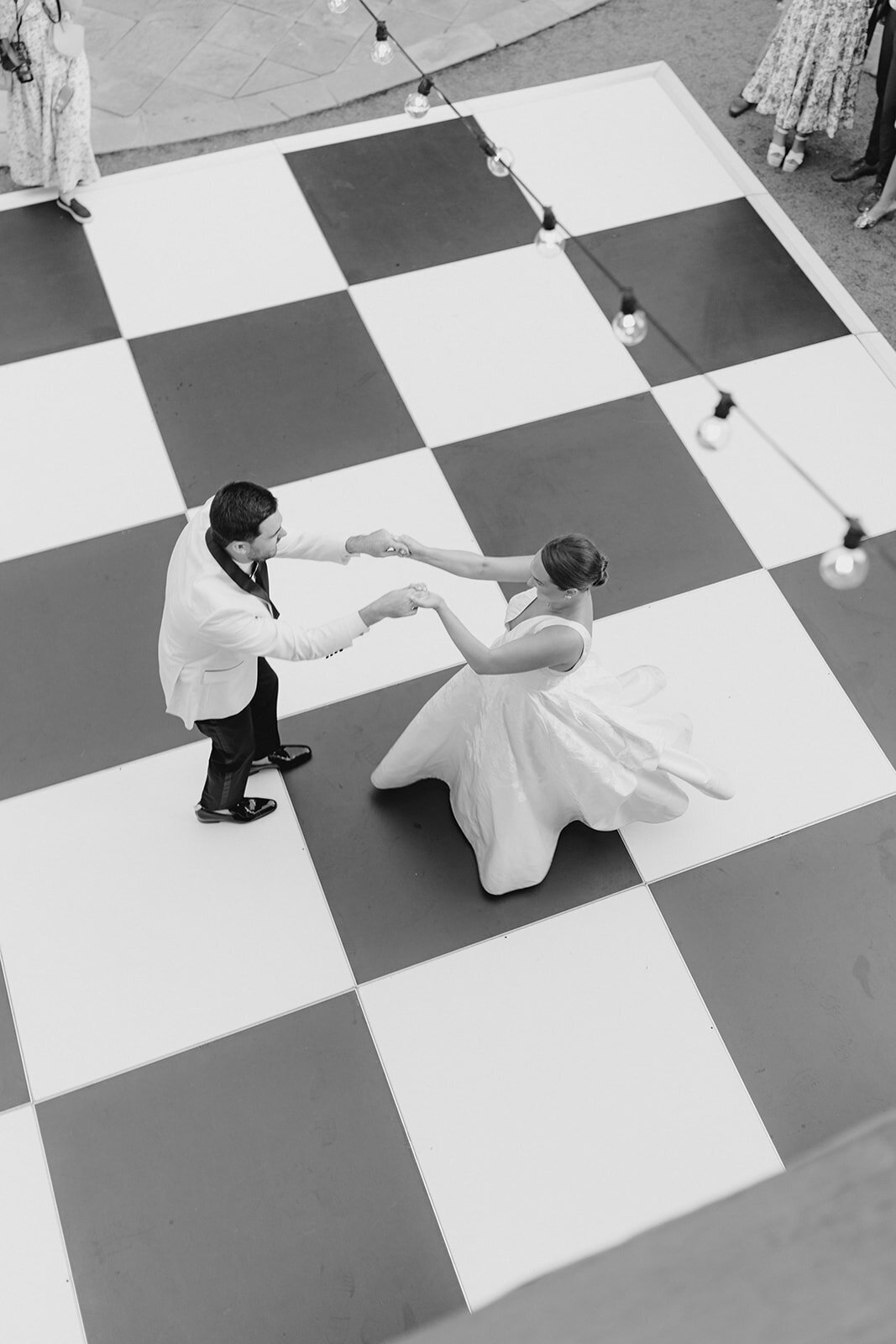 Bride in Anne Barge wedding dress and groom changed into white tuxedo jacket for wedding reception. Black and white photo from above of bride and grooms first dance with checkerboard floor and string lights. Open air wedding reception at William Aiken House. Charleston spring wedding. Black and white destination wedding photographer. Kailee DiMeglio Photography.
