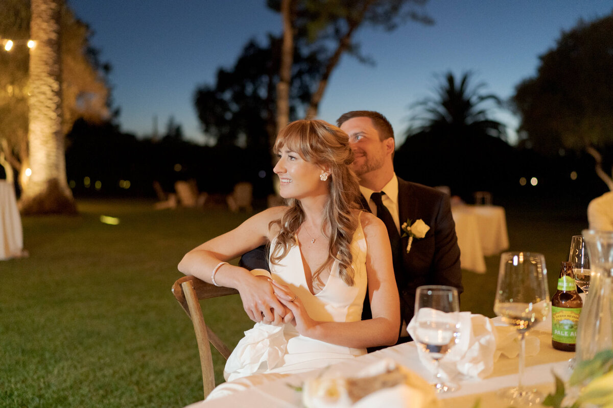 Newly-wed bride and groom sit together and watch the afterparty proceedings in a warmly illuminated evening.
