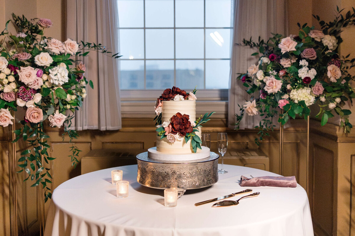 A white wedding cake adorned with burgundy and pink flowers sits on a decorative stand on a round table. Three small candles, a cake server, and a knife are on the table. Floral arrangements and a large window are in the background.