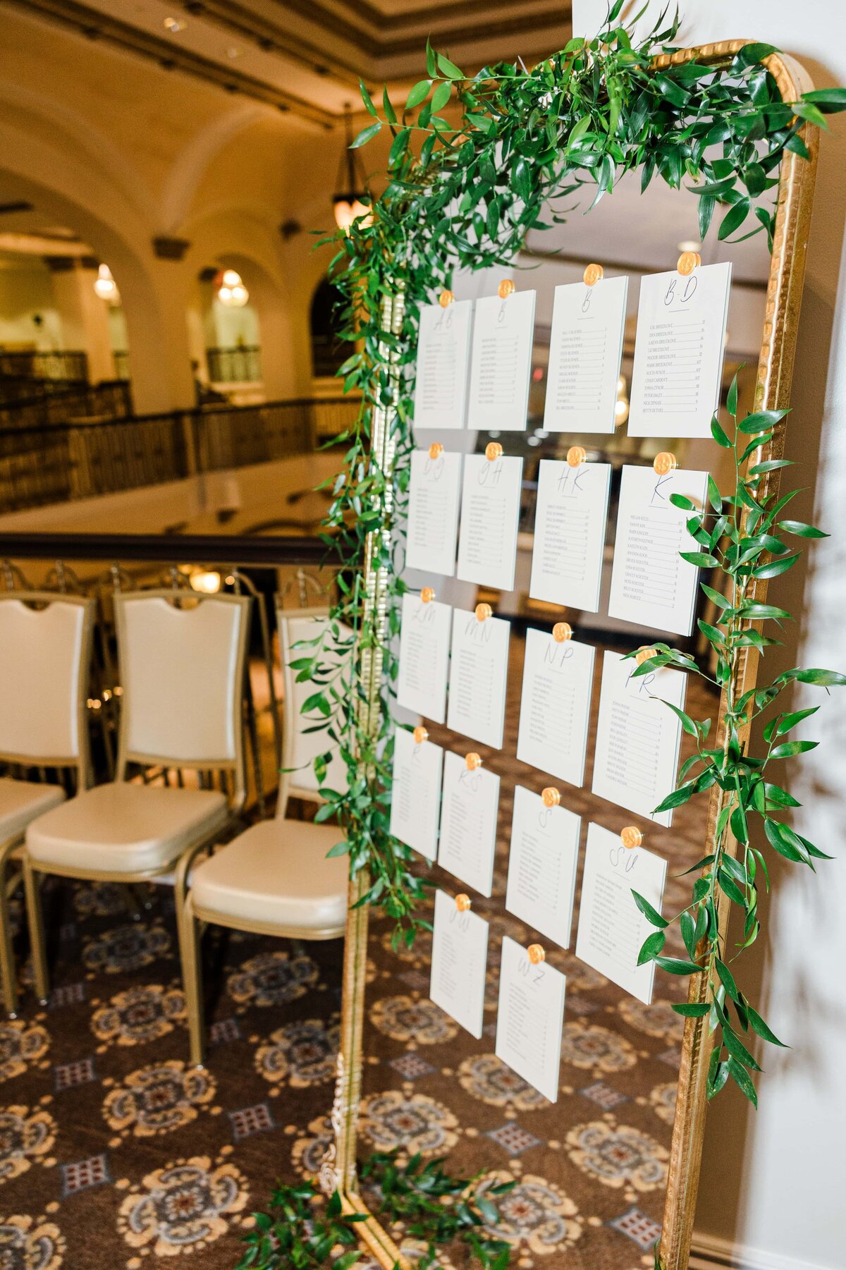 Seating arrangement display at a wedding in Iowa, featuring names listed on white cards hanging from a gold frame adorned with green vines, set in a room with elegant chairs and patterned carpet.