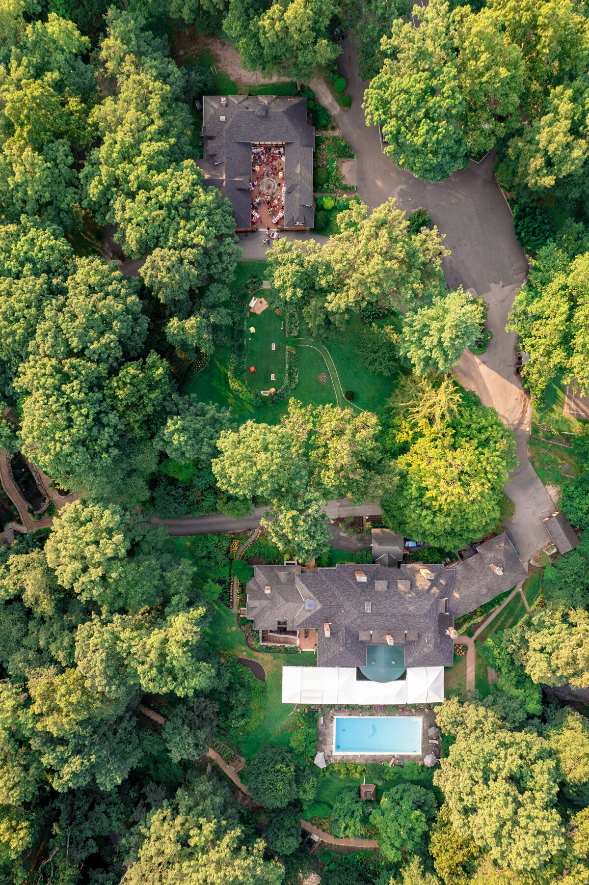 Aerial view of a wooded area with two large houses surrounded by dense trees. One house has a pool and a light-colored canopy. The other house has people gathered on the patio, with paths and greenery throughout the property.
