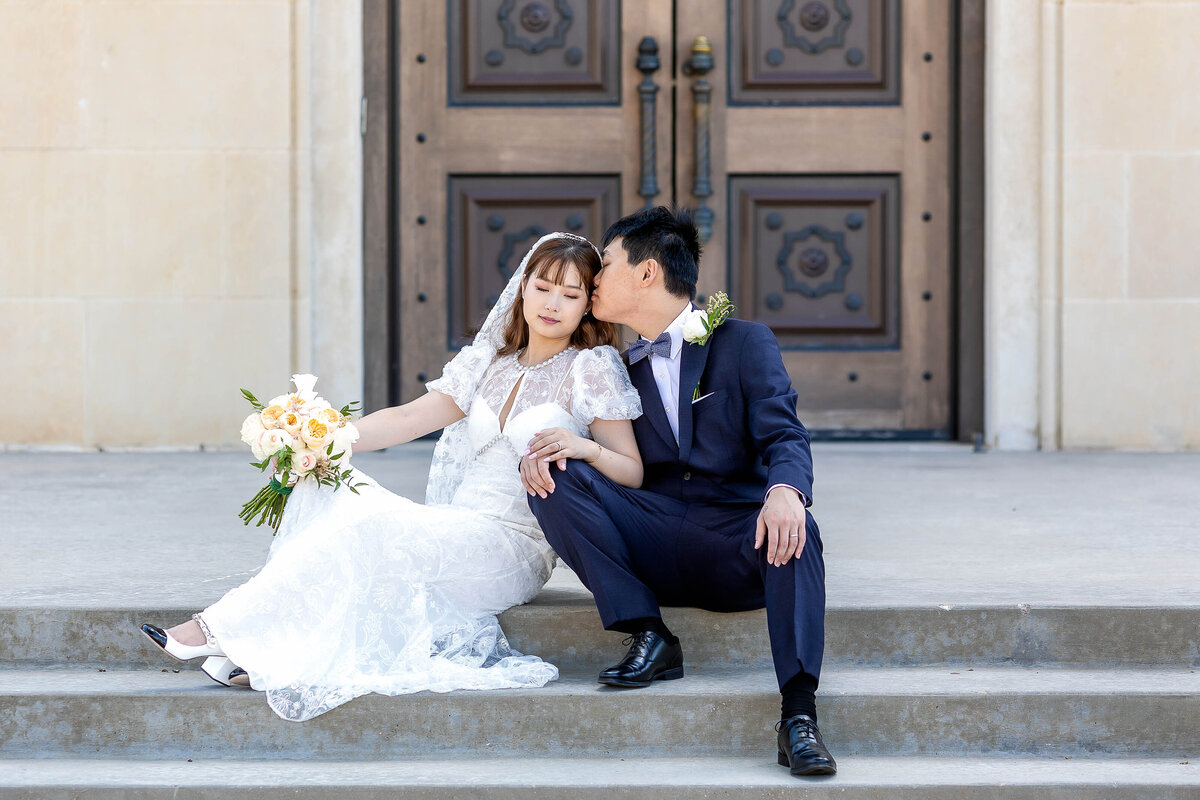 Chinese couple elopement.  Bride and groom are seated on the steps at the Kent Hance Chapel in Lubbock  TX.  Bride is holding her bouquet while leaning into the groom while groom is kissing her on the cheek