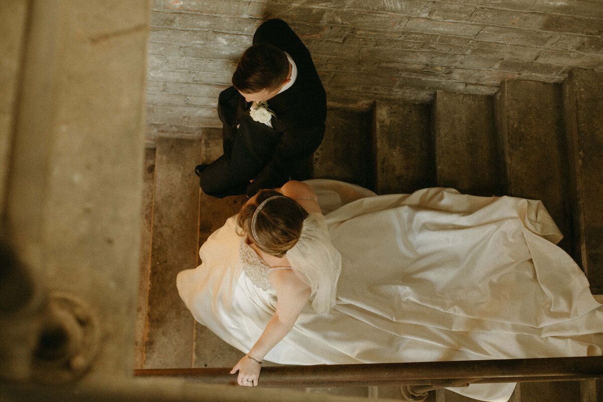 A bride and groom holding hands while descending a concrete stairway in a park, viewed from above. The bride is wearing a long white dress and the groom is in a black suit.