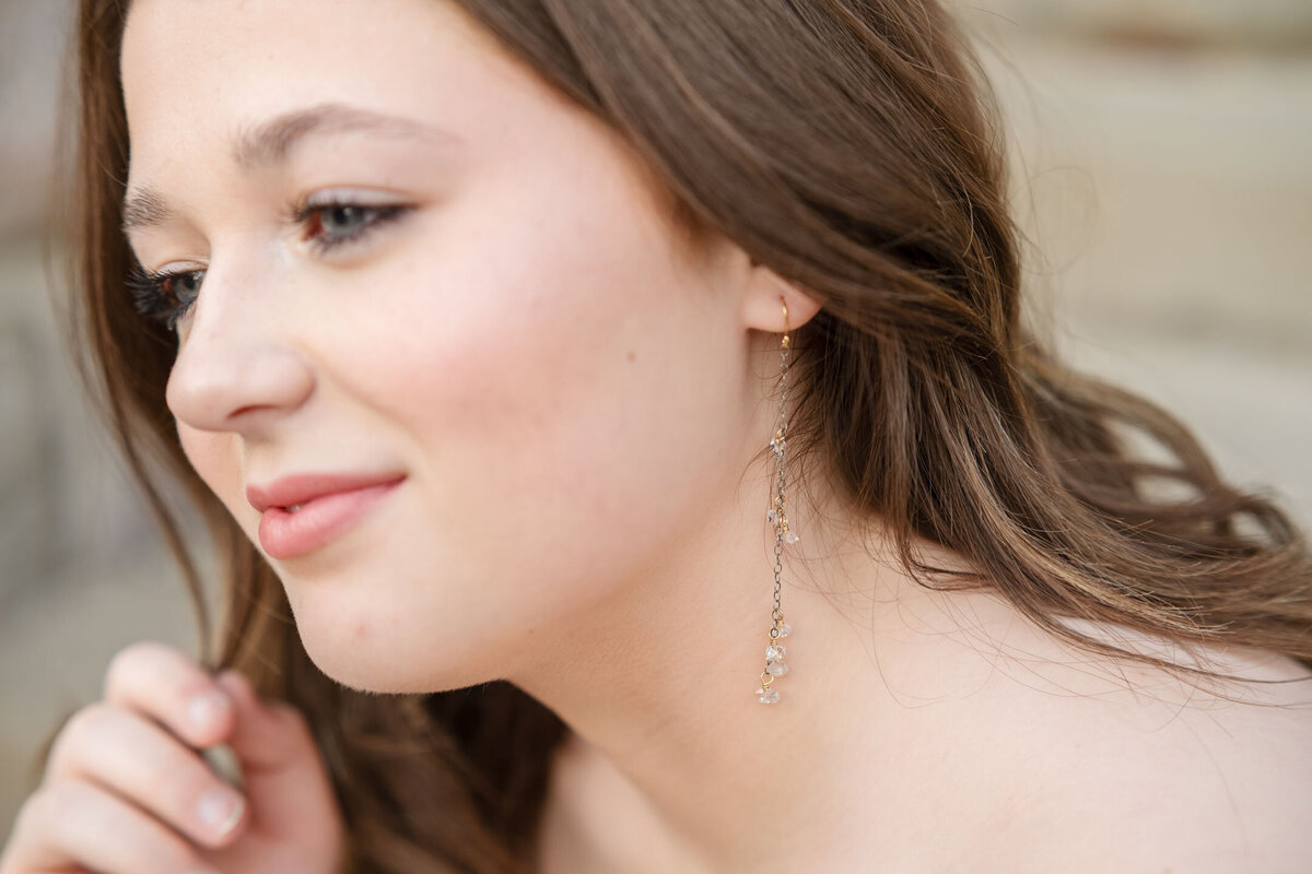 Close-up of a young woman with long brown hair, smiling gently, wearing subtle makeup and dangling earrings.
