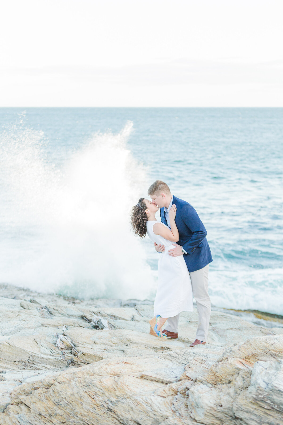 A couple stands on the rocky cliffs of Beavertail Lighthouse for their engagement photos. They have their arms wrapped around each other and share a kiss as the waves crash and create a spray behind them. Captured by best Rhode Island Wedding Photographer Lia Rose Weddings
