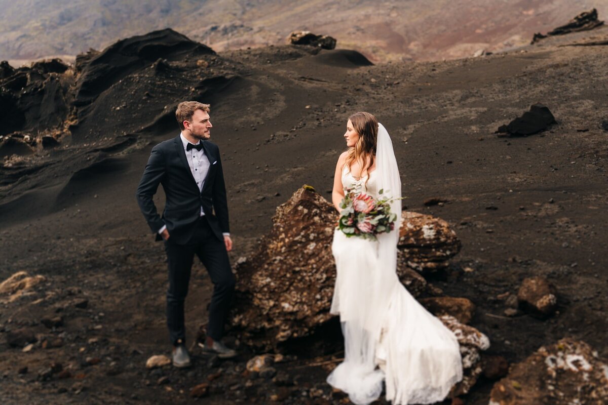 Adventurous couple in Iceland, bride seated on a rock while the groom stands, sharing a heartfelt gaze.