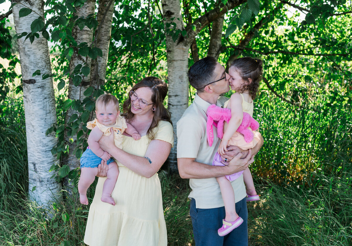 Family in the park with greenery in the back round. Parents looking at the kids and kissing their cheeks.