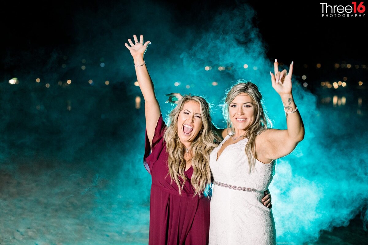 Bride and her Maid of Honor wave to the photographer as they pose with a blue mist behind them