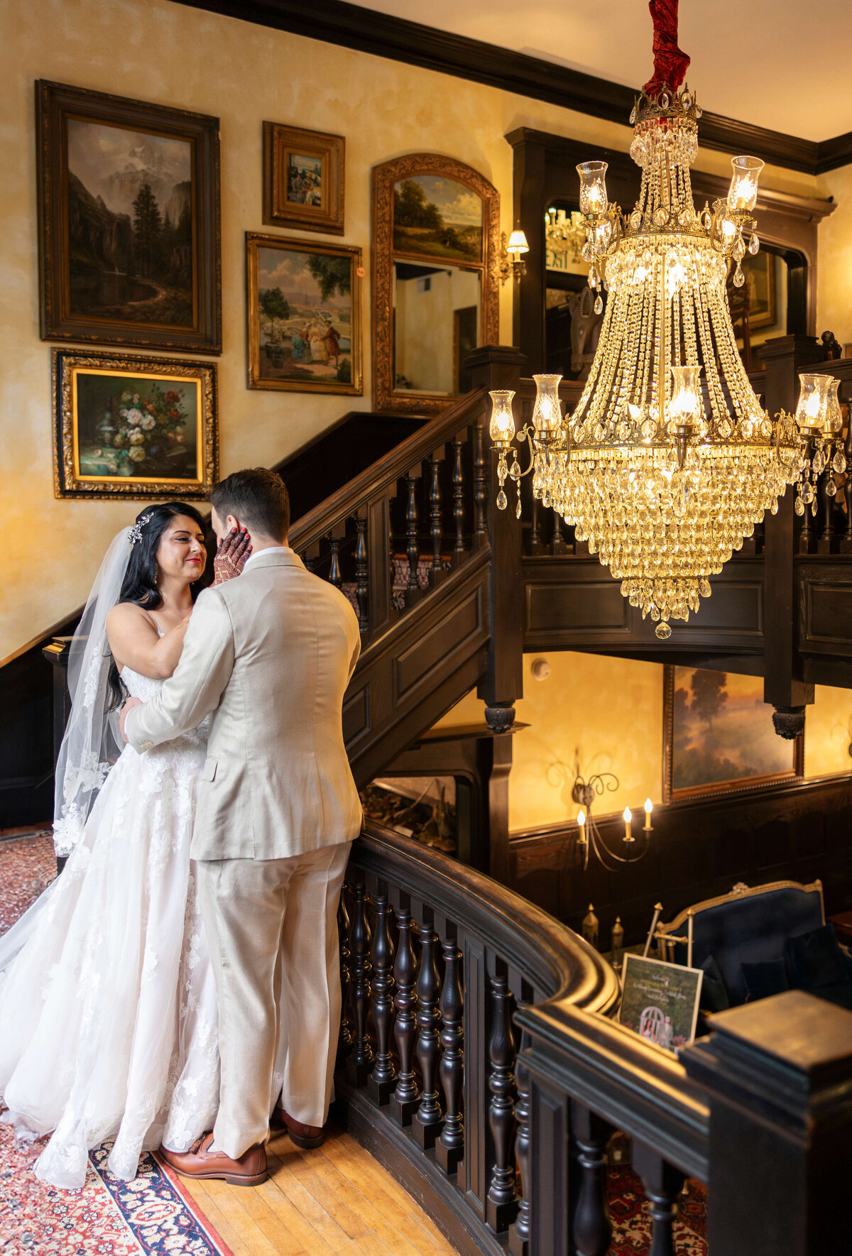 A bride and groom stand on a wooden staircase under an ornate chandelier. The bride is wearing a white wedding dress and veil, while the groom is in a light suit. They are embracing, surrounded by framed paintings on the walls.
