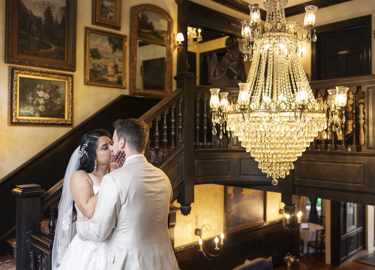 Bride and groom share a moment on a staircase under a sparkling chandelier. The staircase is adorned with framed paintings, creating an elegant atmosphere. The bride is in a white dress and veil, and the groom is in a light-colored suit.